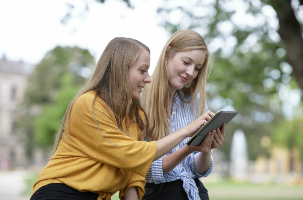 Zwei Studentinnen sitzen draußen und schauen zusammen auf ein Tablet.